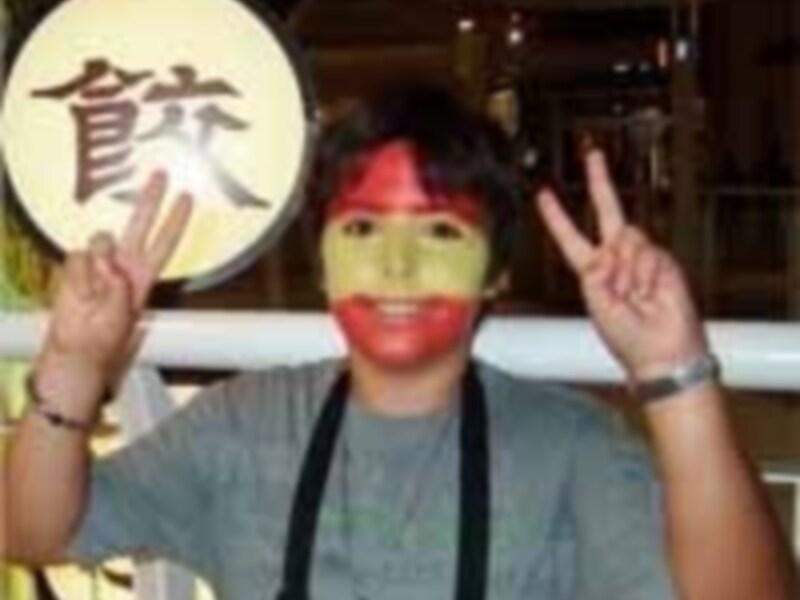 Un joven con una máscara de comida japonesa y un gesto de paz, posando frente a una pancarta con caracteres chinos.