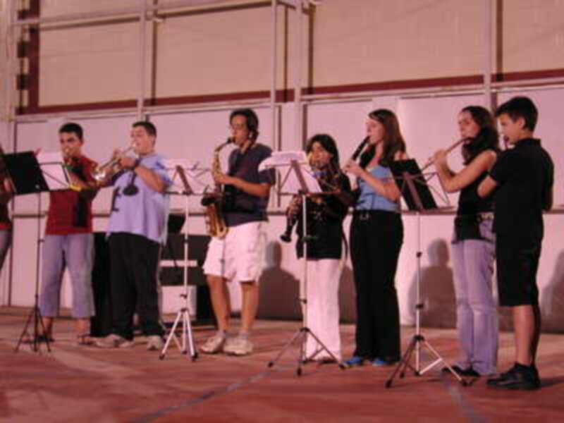 Un grupo de músicos toca instrumentos en una escena al aire libre. El ambiente es festivo y el público está presente, disfrutando de la música.