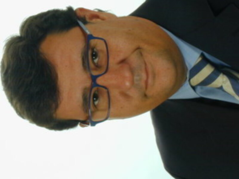 Un hombre con cabello oscuro y gafas azules, vistiendo una camisa blanca y un traje negro.