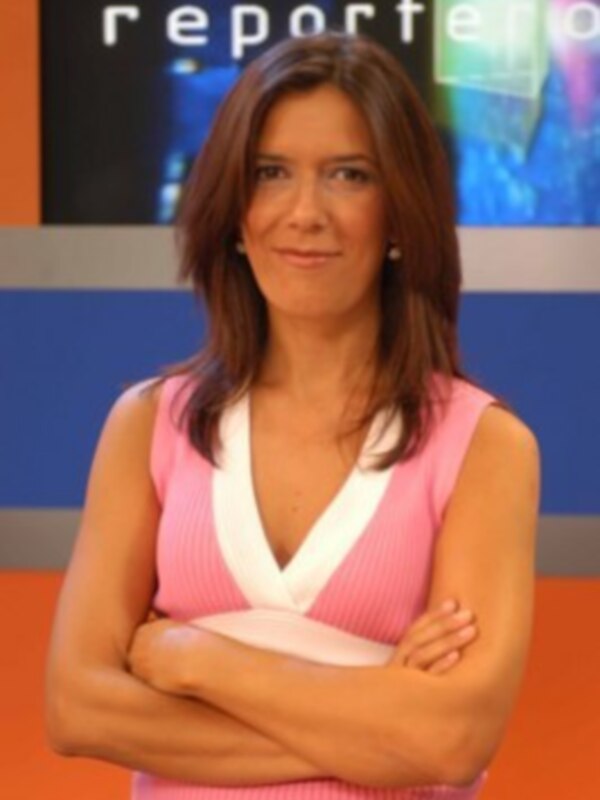 A una mujer con cabello largo y ondulado, vestida con un vestido rosa claro que tiene una escote en V blanco. Está posando con los brazos cruzados y sonriendo frente a un fondo que incluye el texto "reportero" en azul.