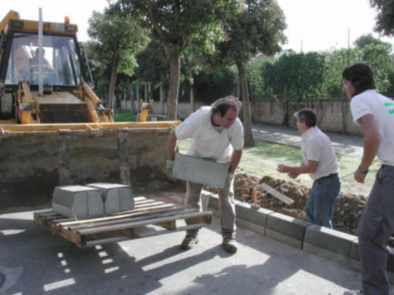 Trabajadores cargando bloques de hormigón en una plataforma, con un tractor excavador al fondo.