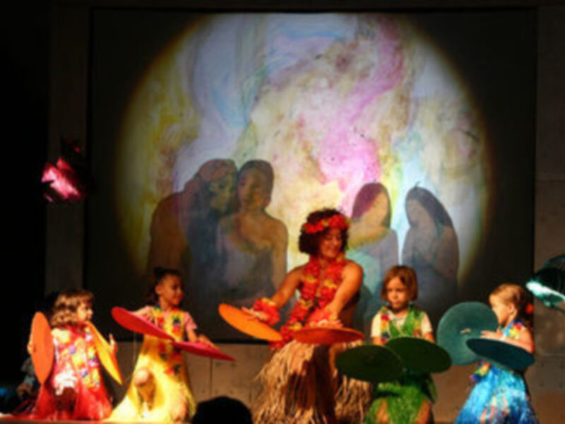 Niños vestidos con disfraces de hula danzan en un escenario frente a una proyección de la Tierra.