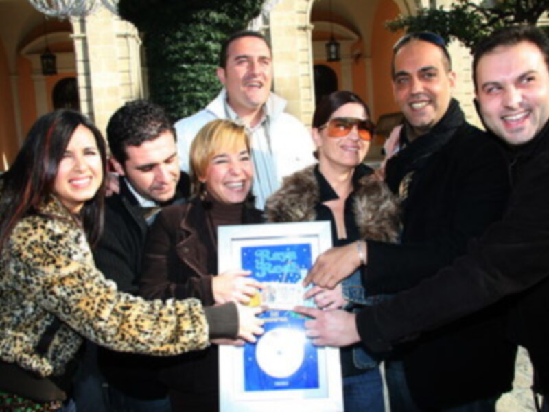 Una grupo de personas sonríe mientras sostienen un certificado. La imagen muestra una celebración o reconocimiento, con algunos portando abrigos y gafas de sol. La escena parece ser al aire libre, frente a un edificio histórico con columnas y ventanas.