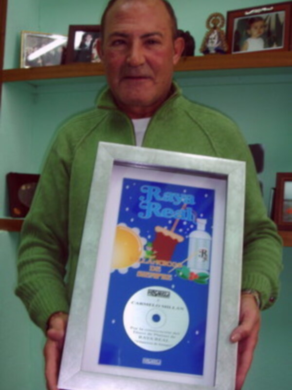 Un hombre sostiene un CD dentro de una marco con un diseño azul y blanco que muestra una imagen de un fruto y el nombre "Raza Real". El fondo es azul con un cielo estrellado.