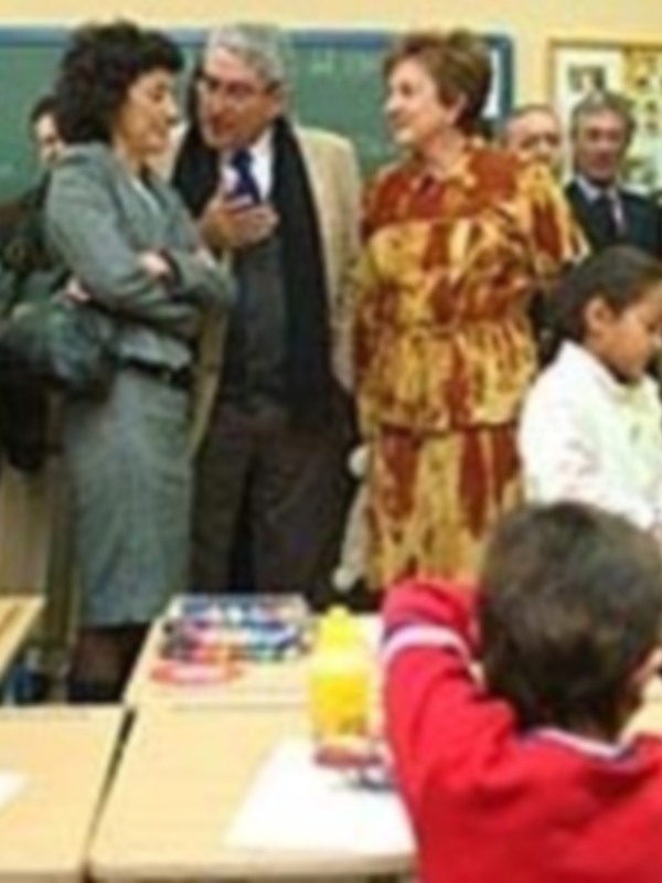 Una escena en una aula con niños y adultos. Un niño en primer plano sostiene un libro, mientras otros estudiantes observan atentos a una persona mayor. Los adultos conversan entre sí en el fondo, y la atmósfera es educativa y familiar.