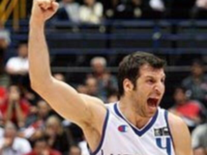 Un jugador de baloncesto celebra emocionado en el estadio, levantando el puño izquierdo. La camiseta lleva el número 4 y la palabra "WOLVERINE". El fondo muestra a otros espectadores y una pantalla de televisión.