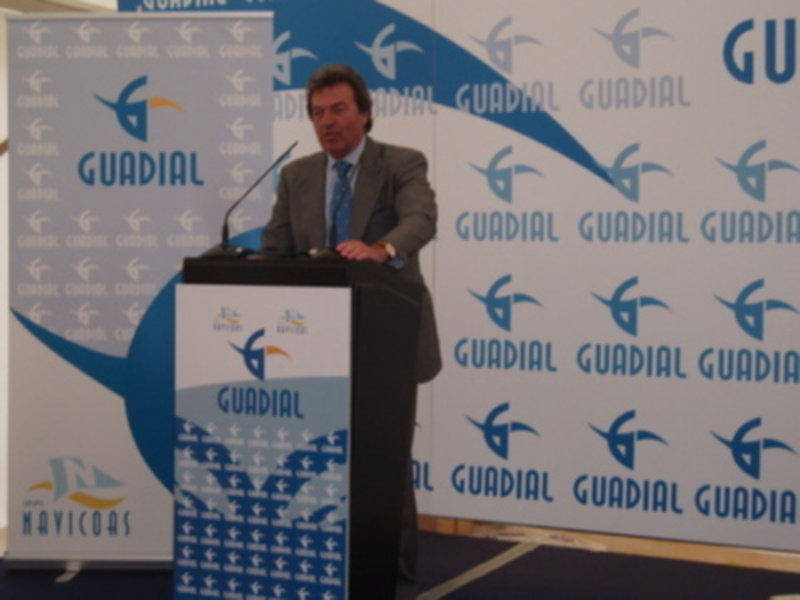 El hombre está en un escenario con un fondo azul y blanco que tiene el logo de Guadal. Está hablando en un micrófono en un podio con el mismo logotipo.