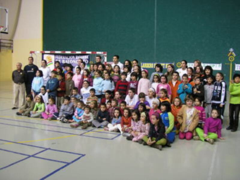 Una escuela primaria celebra un evento de baloncesto con una gran foto de grupo. Los niños y niñas están sentados en el suelo, sonriendo para la cámara. La escuela tiene un baloncesto y una cancha de fútbol al fondo.