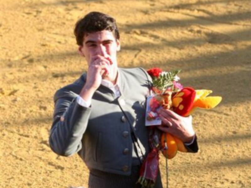 Un joven sostiene una corona de flores y una placa mientras se acerca a besarla. El fondo es un paisaje desértico con sombras de árboles en el suelo.