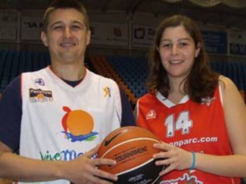 Dos jóvenes posan con un balón de baloncesto en una cancha deportiva. La imagen muestra a una persona con una camiseta blanca y otra con una roja, ambos sonrientes. El fondo muestra gradas vacías y banners de promoción deportiva.