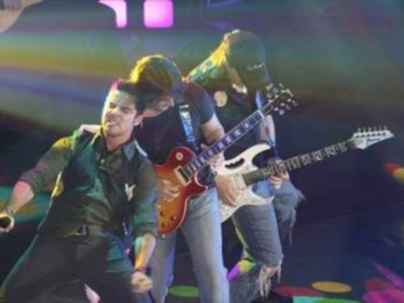 Dos músicos en un escenario iluminado, uno tocando una guitarra eléctrica y el otro con una camiseta verde.