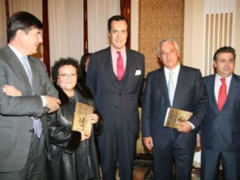 Un grupo de personas en trajes formales sostiene libros en un evento formal, posiblemente una presentación o conferencia.