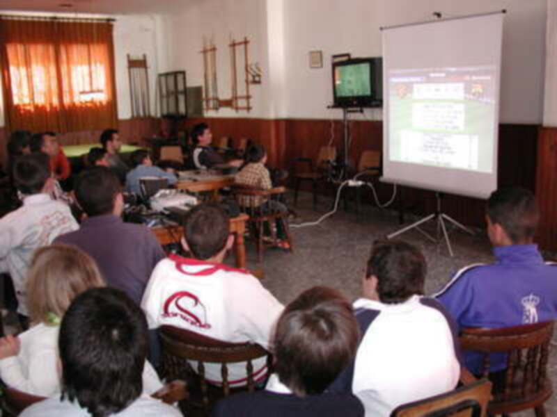 Una clase en una escuela con estudiantes sentados en sillas, viendo un televisor y una proyección de presentación.
