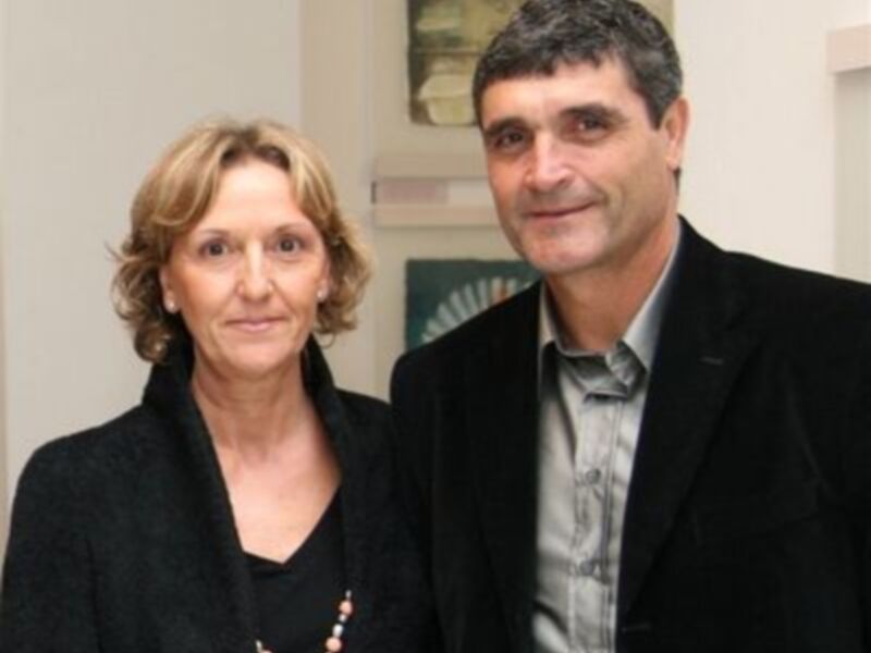 Dos personas sonrientes, uno de ellos con una camisa gris y el otro con una chaqueta negra, posando frente a una pared con un cuadro.