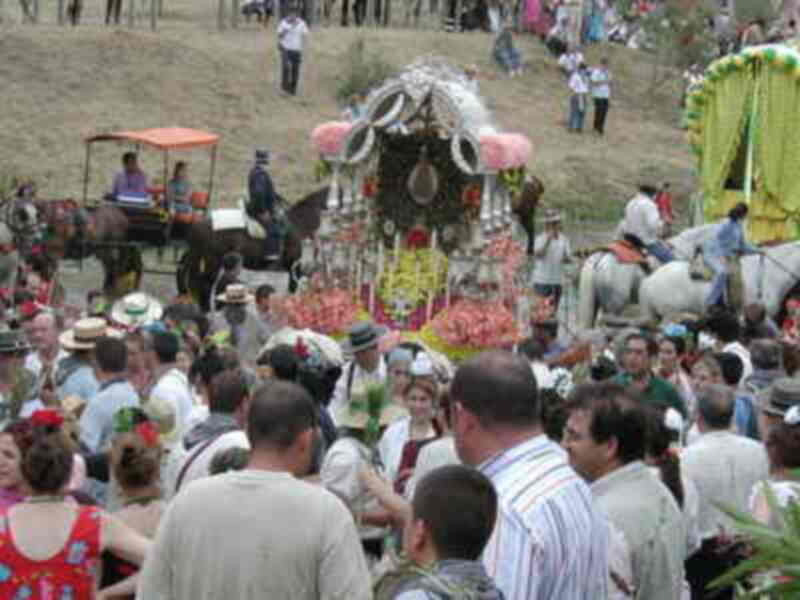 Una multitud de personas celebra una procesión religiosa con carrozas adornadas y jinetes. La escena se desarrolla en un entorno rural con montañas al fondo y una granja cercana.