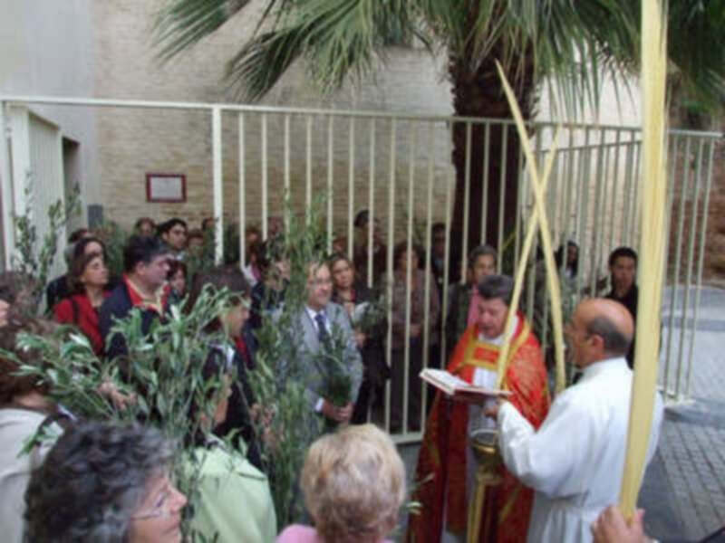 Una procesión religiosa con personas vestidas de rojo y blanco, un sacerdote en mitra, palmas y una iglesia al fondo.