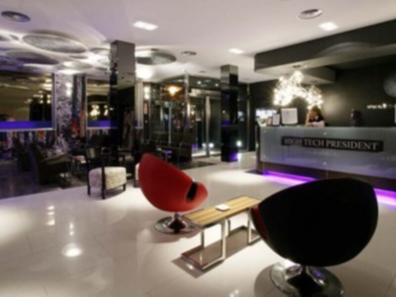 Salón moderno con sillones rojos y negros, mesas de centro, luces colgantes y paredes con letras "HIGH TECH PRESIDENT".