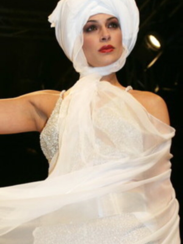 A una modelo con un vestido blanco de encaje y un turbante blanco. La postura elegante y la atención al detalle en el vestuario sugieren un evento de moda o una presentación de diseñadores.