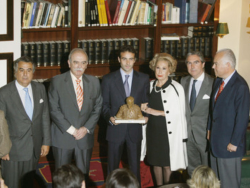 Un grupo de hombres y una mujer en trajes posan para una foto en un salón con estanterías de libros y lámparas. La imagen muestra a los hombres sosteniendo un trofeo mientras la mujer sostiene una placa.