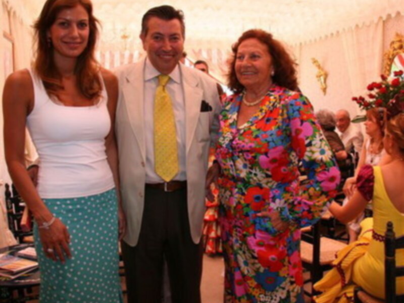 Una imagen festiva de una familia en un evento formal, con una mujer en el centro vestida con un vestido floral vibrante y dos adultos a su lado, uno de ellos con una corbata amarilla.