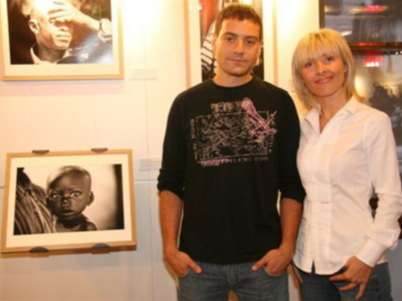 Dos personas posan frente a una exposición de arte, con una imagen en blanco y negro de un niño en el fondo.