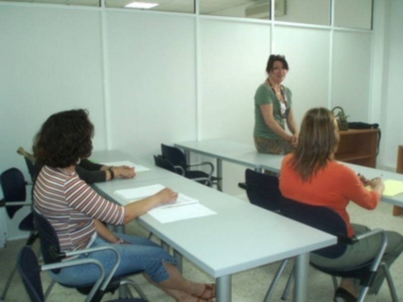 Tres personas sentadas en sillas frente a una mesa, con una mujer de pie detrás. La imagen parece ser de un ambiente educativo o de trabajo en grupo.