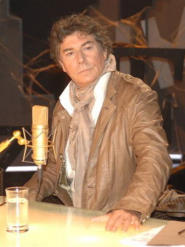 Un hombre con cabello rizado y barba, vestido con una chaqueta marrón y un pañuelo blanco. Está sentado a una mesa con un vidrio de agua en frente y parece estar en un evento o conferencia, con una iluminación dramática y un fondo con luces.