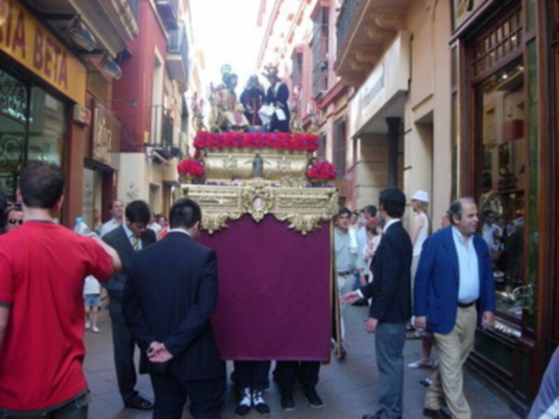 Una procesión religiosa con una imagen de Cristo en un carro, rodeada por personas vestidas formalmente. El entorno muestra edificios antiguos con comercios abiertos y gente caminando.
