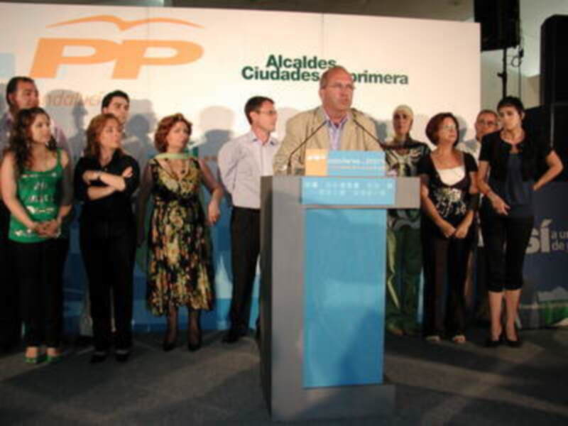 Un hombre habla en un podio con el logo del PP y la frase "Alcaldes Ciudades Primera". A su lado, varias personas escuchan atentamente. La imagen parece ser de un evento político en España.