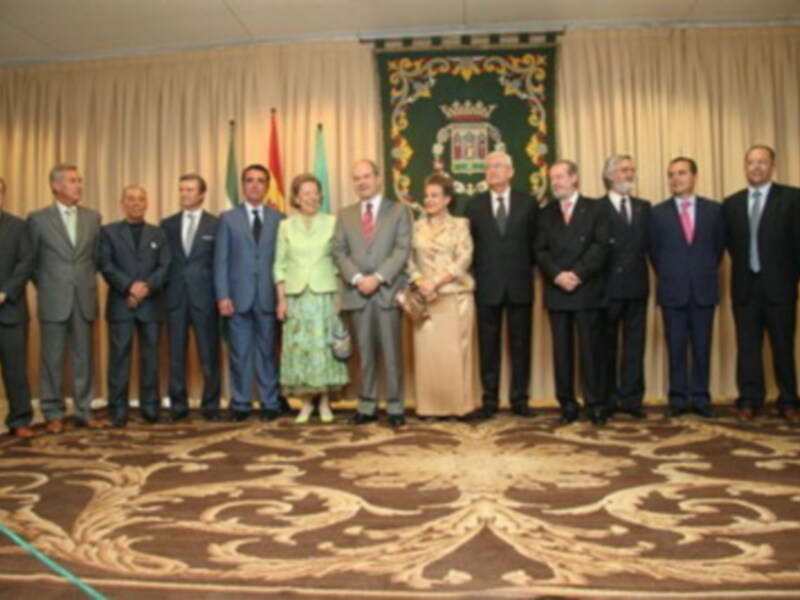Una reunión formal con una fila de personas vestidas con trajes y falda, posando frente a un escudo heráldico.