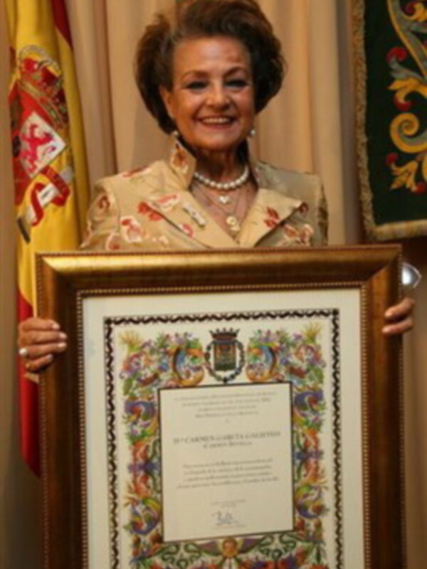 A una mujer sonriente sosteniendo un diploma con un marco elaborado. El fondo incluye una bandera española y otro cuadro decorativo.