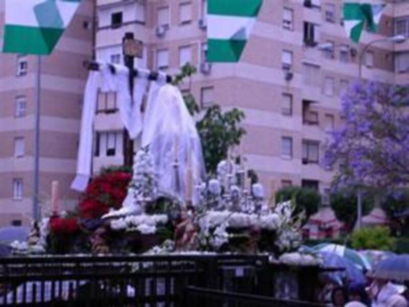 Un trono de madera con una figura central cubierta por mantas blancas, rodeada por flores y velas. En el fondo, edificios con balcones y banderas verdes y blancas ondeando.