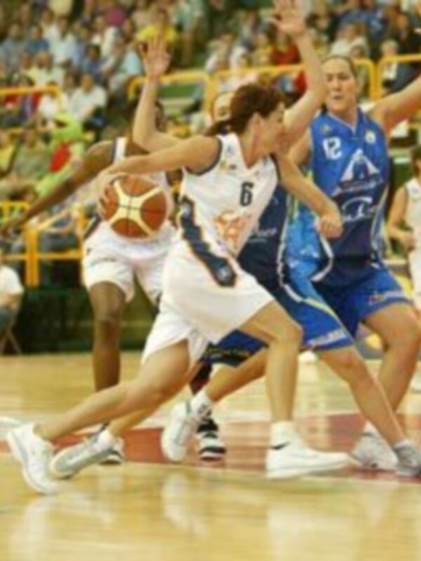 Un jugador de baloncesto blanco intenta bloquear el lanzamiento de un jugador azul durante un partido. El fondo muestra a otros jugadores y espectadores en el estadio.