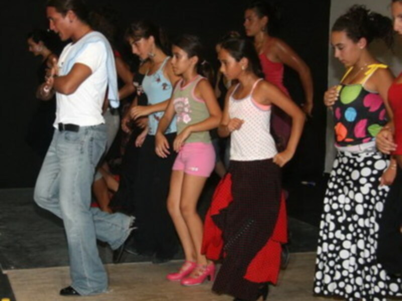 Una escena de una fiesta en la que varias personas están bailando y disfrutando. Se puede ver a una mujer con un vestido rosa y pantalones cortos, y a una mujer con un vestido blanco y pantalones de lunares negros. El ambiente es animado y festivo.