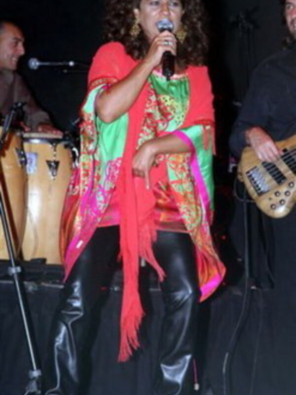 La cantante se encuentra en escenario, con un vestido colorido y una bufanda roja. La guitarra eléctrica de fondo sugiere un concierto en vivo.