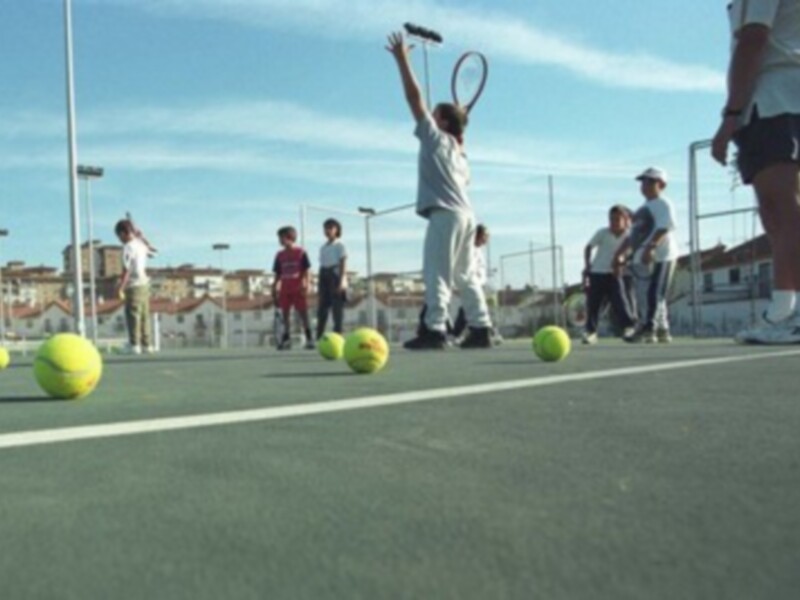 Un niño en un campo de tenis se prepara para servir la pelota, mientras otros niños observan y juegan con pelotas de tenis en el suelo. La escena se desarrolla bajo un cielo azul claro, con edificios y una cerca en el fondo.