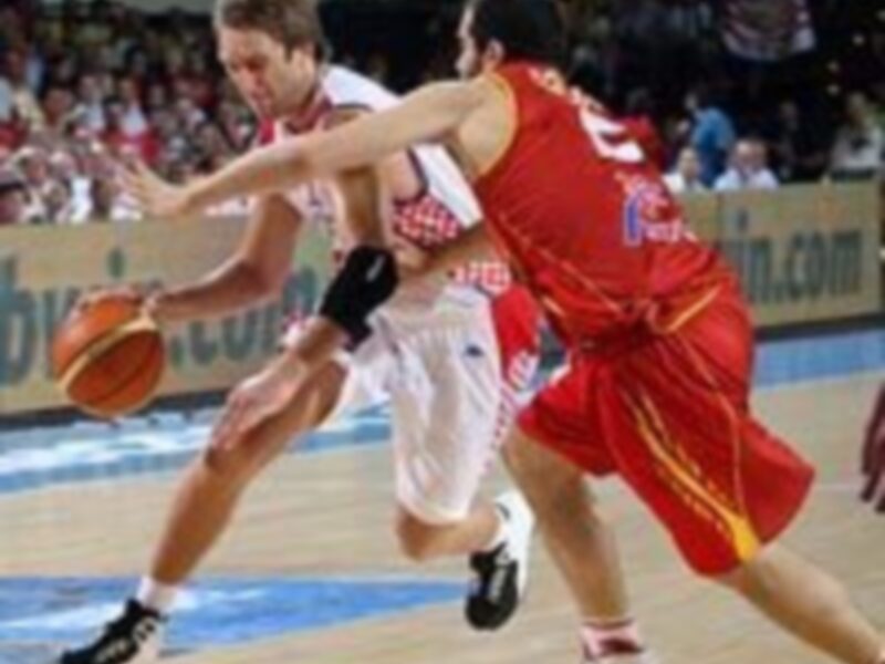 Juego de baloncesto en un estadio lleno de espectadores, con dos jugadores en acción, uno con camiseta roja y otro con camiseta blanca con detalles rojos.