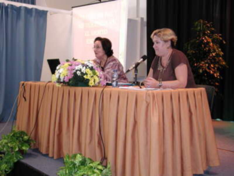 Dos mujeres sentadas en una mesa con un fondo de cortinas y una pantalla. La mesa está cubierta con una manta naranja, hay flores en el centro y un microfono en la mesa. La imagen parece ser de una conferencia o evento profesional.
