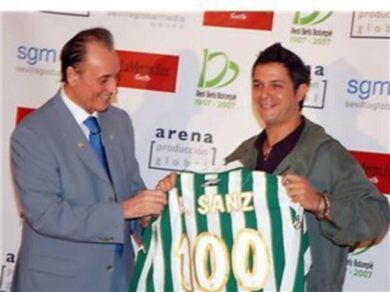 Un hombre en traje de negocios y otro en camisa verde con el número 10, posando con una camiseta de fútbol en un evento de productos.