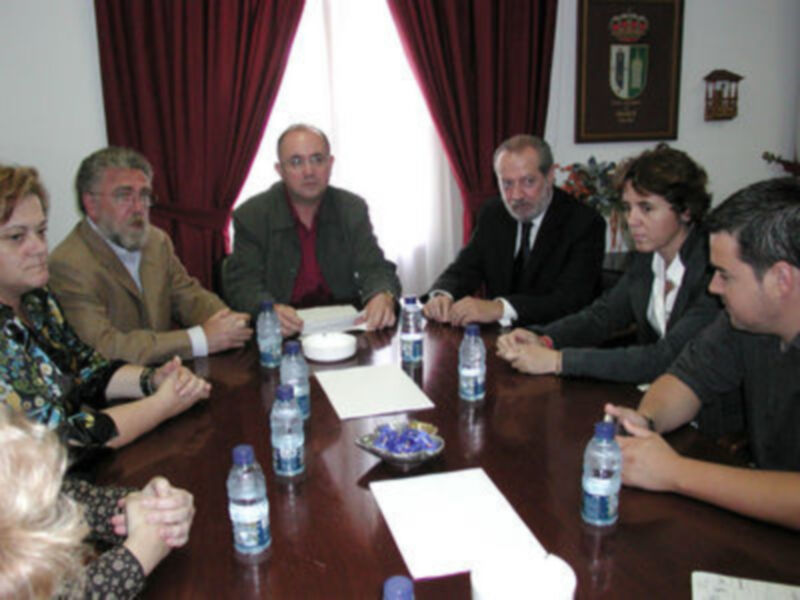 Una reunión formal en una sala con cortinas rojas y un escudo en la pared. Personas sentadas alrededor de una mesa con botellas de agua y documentos.