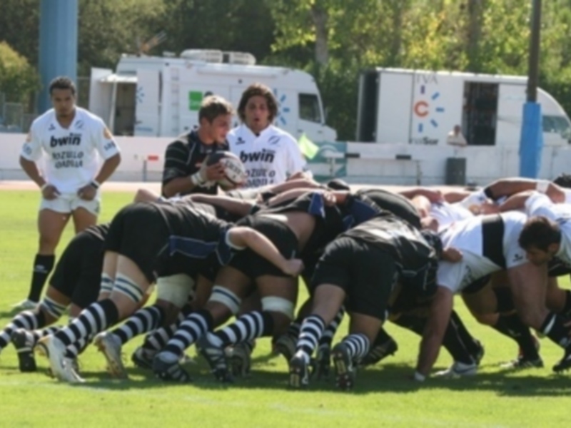 Un grupo de jugadores de rugby en una formación de bloque en un campo de juego verde, con camisetas blancas y negras, y una camioneta blanca en el fondo.