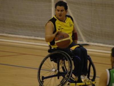 Un hombre en silla de ruedas juega baloncesto. El fondo es una cancha deportiva con un objetivo de portería visible.