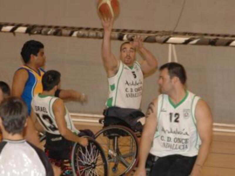 Un jugador en silla de ruedas sostiene un balón mientras otros jugadores lo observan durante un partido de baloncesto.