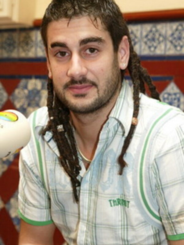 Un hombre con trenzas negras y un barba corta, vistiendo una camisa a cuadros verde y blanco. Detrás de él se observa un muro con azulejos de diseño geométrico.