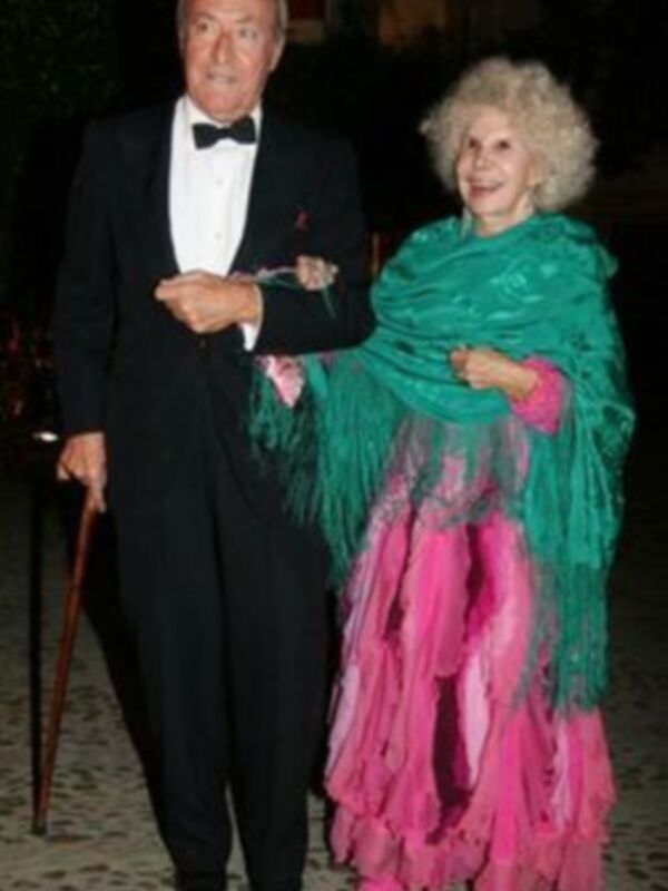 Un hombre en traje negro y corbata, posando junto a una mujer con un vestido rosa y verde. Ambos están en un ambiente formal, posiblemente una fiesta o evento.