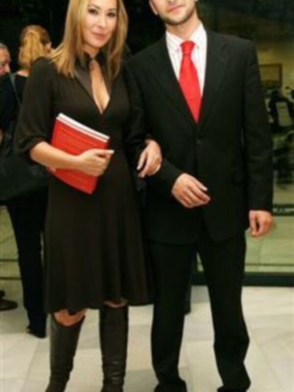 Un hombre y una mujer posando juntos, el hombre lleva traje y corbata roja, la mujer lleva vestido negro y botas altas.