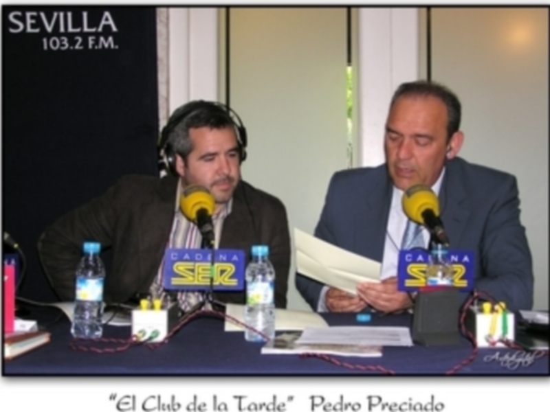 Dos hombres en un estudio de radio, uno con auriculares y el otro sosteniendo un papel. En la mesa hay botellas de agua, micrófonos y una bandera con "SEZ 103.2 F.M." y "El Club de la Tarde" Pedro Preciado.