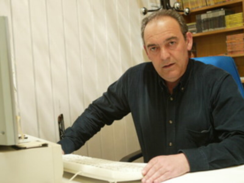 Un hombre con una camisa azul está sentado frente a un escritorio, usando un teclado y mirando la cámara. En el fondo, hay una estantería con libros y un monitor de computadora.