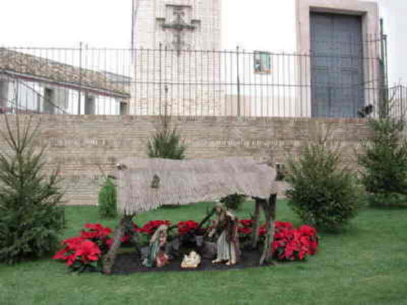 Un belén navideño en un jardín frente a una iglesia con una cruz visible. La escena está rodeada de plantas y flores rojas, creando un ambiente festivo.