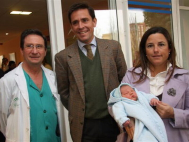En una clínica, un médico en blanco y un hombre con traje sonríen junto a una mujer y su recién nacido en una manta azul.